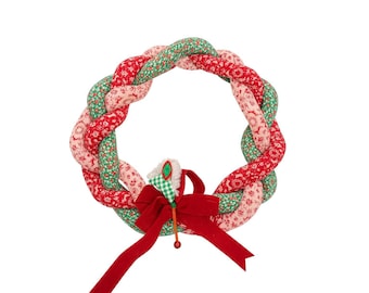 May include: A circular fabric wreath with a braided design, featuring red, pink, and green floral patterns. A red velvet bow and a small decorative element with a green and white checkered pattern adorn the wreath.