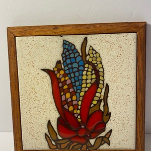 Puede incluir: Un azulejo de cerámica enmarcado con un diseño de mosaico colorido de una mazorca de maíz en tonos rojos, amarillos, azules y marrones. El azulejo tiene un fondo blanco con motas marrones y está enmarcado en un marco de madera oscura.