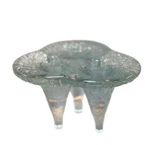 Puede incluir: Un cuenco decorativo de vidrio transparente con tres patas. El cuenco tiene una superficie texturizada y una forma redondeada, similar a una nube. Las patas son cónicas y tienen un ligero tono dorado en la base. El cuenco probablemente se utiliza con fines decorativos.