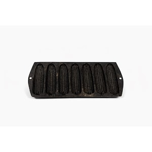 May include: A cast iron cornbread pan with seven corn cob molds. The pan is dark brown and has a rectangular shape with rounded ends. The molds are detailed to resemble corn cobs.