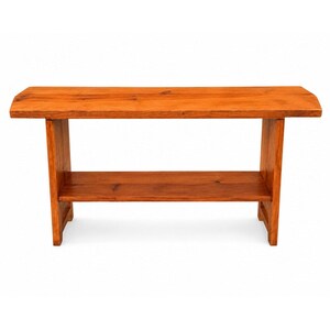 May include: A rectangular wooden bench featuring a warm, medium-brown finish with visible wood grain patterns. The bench is constructed with a flat top surface, two sturdy vertical side supports, and a single lower shelf positioned between the legs.