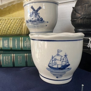 May include: Two white ceramic planters with blue and white designs. One planter features a windmill and the other features a sailboat.