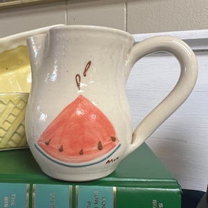 May include: A white ceramic pitcher with a watermelon design. The pitcher has a handle and a spout. The watermelon is red with black seeds and a green rind.