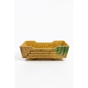 Könnte beinhalten: Eine rechteckige Keramikschale mit strukturiertem, gesprenkeltem gelbbraunem Innenraum und einer grünen Ecke. Die Schale hat ein abgestuftes Design an den Seiten und steht auf kleinen Füßen. Die Gesamtästhetik ist vintage und dekorativ.