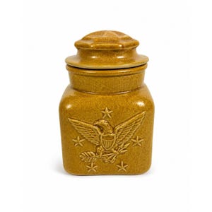 May include: Mustard-colored ceramic jar with a lid. The jar features an embossed eagle emblem with stars. The lid has a star-shaped design. The jar is a square shape with rounded corners.