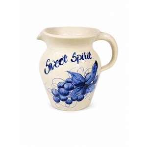May include: A cream-colored ceramic pitcher featuring a blue hand-painted design of grapes and leaves. The text "Sweet Spirit" is written in blue script above the grape illustration. The pitcher has a curved handle and a small pouring spout.