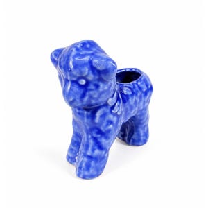 May include: A small, glossy, cobalt blue ceramic figurine shaped like a lamb. The surface features a textured, mottled finish with varying shades of blue. The piece has a hollow opening on the back, suggesting it functions as a small planter or holder.