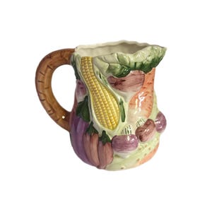 Puede incluir: Una jarra de cerámica con un asa marrón y un diseño de verduras. La jarra presenta una mazorca de maíz, berenjenas, zanahorias y otras verduras en tonos de amarillo, morado, naranja y verde. La jarra tiene un asa texturizada y un interior blanco.