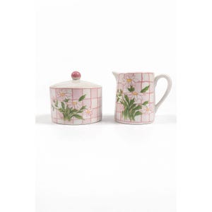 May include: A ceramic sugar bowl with a lid and a creamer pitcher. Both are decorated with a pink grid pattern and white daisy floral designs. The sugar bowl has a pink knob on the lid.