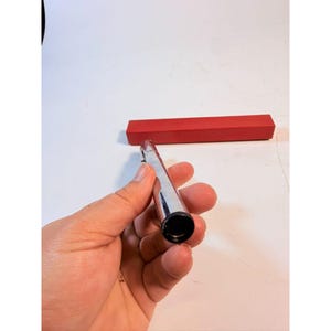 May include: A silver-colored, cylindrical object with a black opening is held in a hand. A red rectangular box is in the background. The object appears to be a pen or a similar writing instrument.