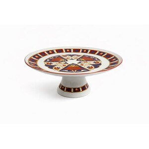 May include: A white ceramic cake stand with a decorative design. The plate has a red border and a pattern of red, blue, and gold shapes. The base of the stand has a matching design. A classic piece for serving desserts.