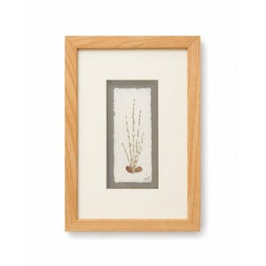 May include: A framed artwork featuring a botanical arrangement. The artwork showcases a vertical composition of dried stems and seed pods on textured paper, set against a gray background. The artwork is encased in a light wood frame with a white mat.