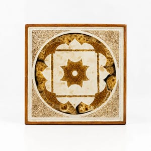 May include: Square decorative tile with a geometric design. The tile features a central star shape and a surrounding circular pattern in shades of brown and cream. The tile is framed by a wooden border.