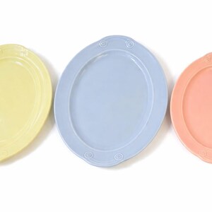 Puede incluir: Tres fuentes ovaladas para servir en colores pastel: amarillo, azul claro y melocotón. Cada fuente tiene un borde elevado y detalles decorativos en los extremos. Las fuentes están dispuestas sobre una superficie blanca.