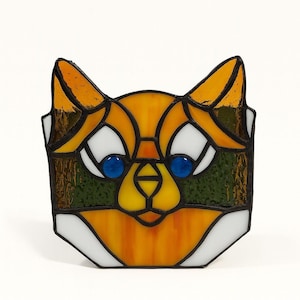 Op de afbeelding: Gebrandschilderd glazen vossenkop met oranje, geel, wit, groen en blauw. De vos heeft grote witte ogen met blauwe pupillen, oranje oren en een gele neus. Het glas is omlijst met een donkere rand.