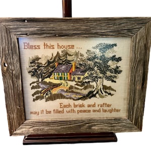 May include: A framed cross-stitch with a rustic wooden frame. The cross-stitch depicts a small house with a bridge and trees. The text reads "Bless this house... Each brick and rafter may it be filled with peace and laughter."