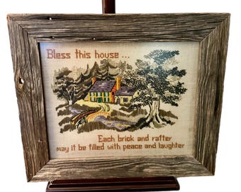 May include: A framed cross-stitch with a rustic wooden frame. The cross-stitch depicts a small house with a bridge and trees. The text reads "Bless this house... Each brick and rafter may it be filled with peace and laughter."