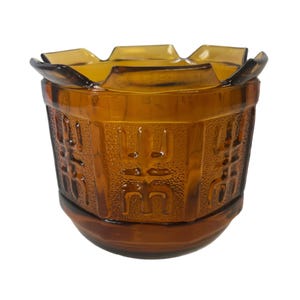 Puede incluir: Jarrón de vidrio ámbar con borde festoneado y caracteres chinos en relieve. El jarrón tiene una superficie texturizada y un fondo redondeado.