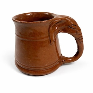 May include: Brown ceramic mug with an elephant head handle. The mug has a cylindrical body with horizontal grooves and a wide opening. The handle is shaped like an elephant's trunk and head, adding a unique design element to the drinkware.