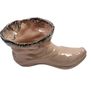 May include: A ceramic boot-shaped planter in a soft pink hue. The rim is accented with a dark, speckled pattern. The boot has a glossy finish and is designed with a rounded toe and a detailed ankle.