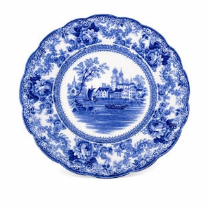 May include: A round, white ceramic plate with a blue floral and landscape design. The plate features a scalloped edge and a central scene of a town by a river, with boats and buildings. The border is decorated with floral patterns.