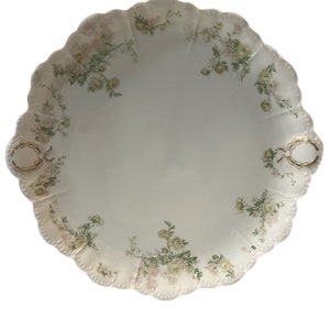 May include: A white porcelain serving platter with a scalloped edge and a floral design in shades of pink and green. The platter has two gold handles.