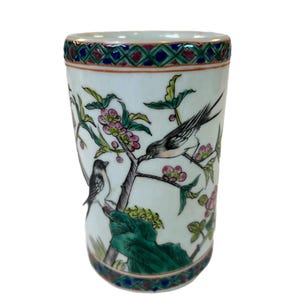 Könnte beinhalten: Ein weißer Keramikbecher mit detailliertem Blumen- und Vogeldesign. Der Becher hat einen dekorativen Rand mit einem geometrischen Muster in Rot, Blau und Grün. Das Kunstwerk zeigt Vögel, die auf blühenden Zweigen sitzen.