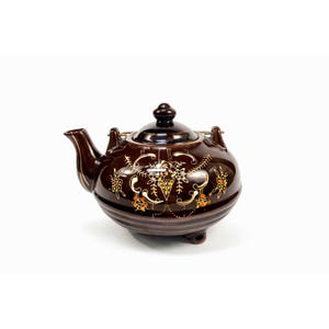 May include: A brown ceramic teapot with a lid and a handle. The teapot has a decorative gold and orange floral pattern. The teapot is on a white background. The teapot has a spout and two small feet.