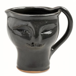 Könnte beinhalten: Eine schwarze Keramikkrug mit einem Gesichtsdesign. Der Krug hat einen Henkel und einen Ausguss. Das Gesicht hat geschlossene Augen und einen kleinen, gepunkteten Mund. Der Boden des Kruges ist naturfarben.
