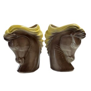 Puede incluir: Dos esculturas de cerámica con forma de cabeza de caballo. Los caballos son marrones con crines amarillas. Las esculturas miran en direcciones opuestas y están sobre un fondo blanco. Probablemente sean objetos decorativos.