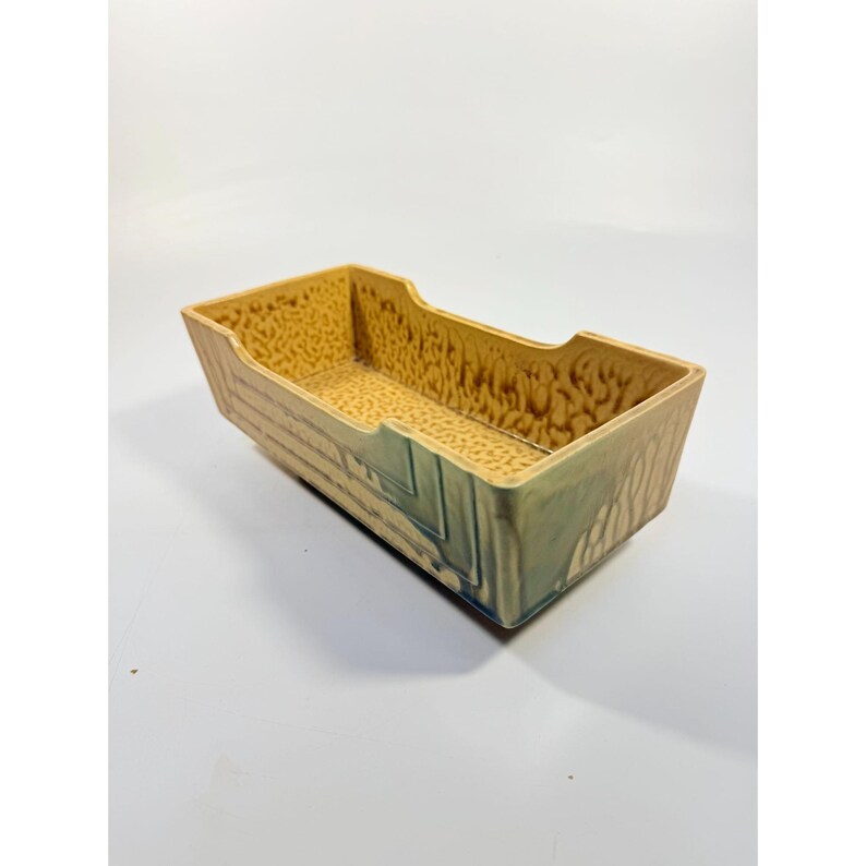 K&ouml;nnte beinhalten: Rechteckige Keramikschale mit strukturierter Innenseite und geometrischem Design. Die Schale hat eine beige-braune Farbgebung mit Akzenten in Blau und Gr&uuml;n. Die Seiten sind abgewinkelt und die Oberseite hat ausgeschnittene Griffe.