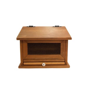 Puede incluir: Una panera de madera con tapa con bisagras y un panel frontal de malla metálica. La panera tiene un pequeño cajón con un pomo blanco. La madera tiene un acabado natural y la panera es rectangular.