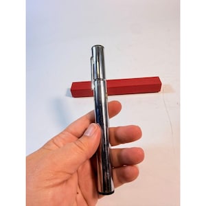 May include: A shiny, silver pen with a pocket clip is held in a hand. A red rectangular box is in the background. The pen is cylindrical and appears to be made of metal. The background is white.