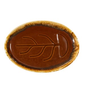 May include: Oval-shaped brown ceramic dish with a leaf design. The dish has a glossy, dark brown interior and a lighter brown, textured rim. The leaf design is etched into the center of the dish.