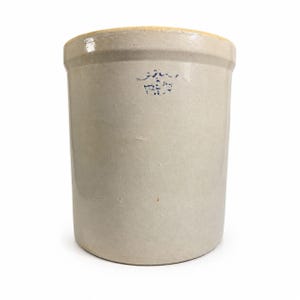 Puede incluir: Un tarro de gres con forma cilíndrica y un esmalte gris claro. El borde es de color beige claro. Un sello azul es visible en la parte delantera. Este artículo de cocina vintage es adecuado para almacenamiento o exhibición.