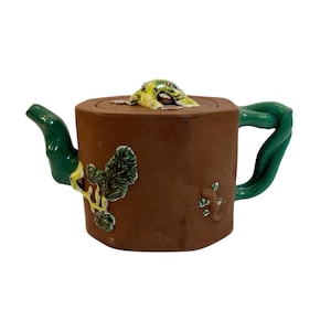 Op de afbeelding: Een bruine keramische theepot met een groene handgreep en tuit. Het deksel heeft een kikkerknop. Groene en gele bloemaccenten versieren de zijkant van de theepot. De theepot is ontworpen met een unieke, artistieke stijl.