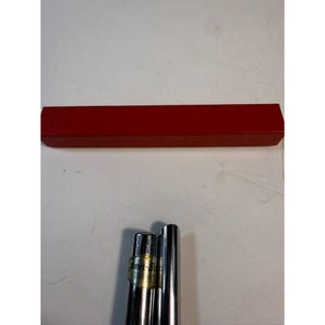 May include: A red rectangular box and three silver metal pen components. The box is a solid, vibrant red. The pen components are cylindrical and shiny, with one featuring a gold and white label.