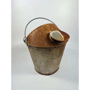 May include: A weathered metal bucket with a rusty brown top and a silver handle. The bucket has a spout and a small, round opening. The body of the bucket is a faded silver color.