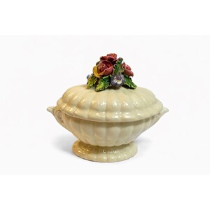 May include: A cream-colored ceramic lidded serving dish with a pedestal base. The lid is adorned with a cluster of colorful ceramic flowers. The dish has a ribbed texture and small handles on either side.