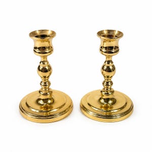 May include: Two gold-colored candle holders with a classic design. Each holder has a round base, a decorative stem with a ball-shaped accent, and a cup-shaped top. The candle holders are made of metal and have a shiny finish.