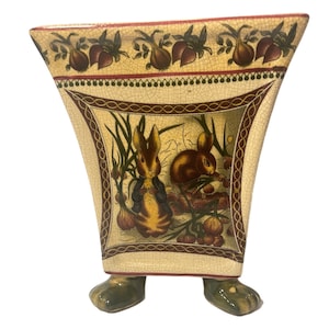 Puede incluir: Un macetero de cerámica con forma cuadrada y cuatro patas. El macetero está decorado con un diseño floral marrón y verde y dos conejos. El macetero tiene un acabado de esmalte craquelado.