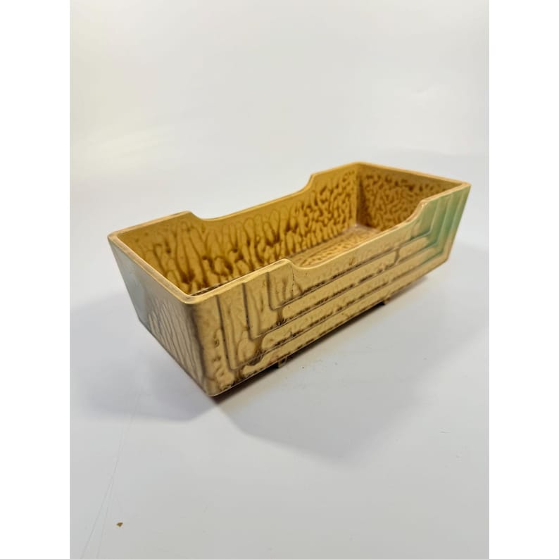 K&ouml;nnte beinhalten: Ein rechteckiger Keramik-Pflanztopf mit strukturiertem Holzmaserungsdesign. Der Pflanztopf ist haupts&auml;chlich beige mit gr&uuml;nen Akzenten an den Kanten. Der Innenraum hat eine strukturierte Oberfl&auml;che. Der Pflanztopf ist leer.