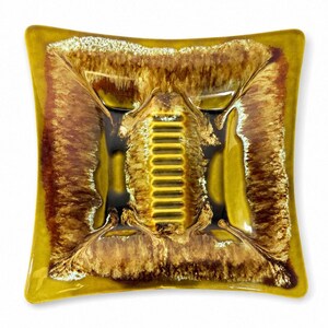 May include: Square ceramic ashtray with a unique design. The ashtray features a combination of brown, gold, and black colors. The center has a raised, textured area with a series of parallel lines. The edges are slightly curved, and the overall shape is square.