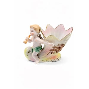 Pode incluir: Estatueta de porcelana vintage com um querubim tocando flauta, sentado em um vaso em forma de concha. O vaso tem uma borda rosa e dourada, com detalhes florais marrons. O querubim usa um tapa-sexo verde.