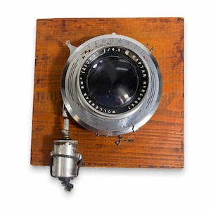 Op de afbeelding: Vintage cameralens gemonteerd op een houten plank. De zilveren lens heeft zwarte tekst, waaronder "RAPTAR" en "WOLLENSAK ROCHESTER U.S.A.". Het heeft metingen zoals "210MM" en "f/4.5". Een metalen component hangt eronder.
