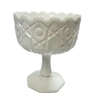 Pode incluir: Uma tigela de pedestal de vidro de leite branco com uma borda festonada e um padrão geométrico. A tigela está sobre um pedestal branco com uma base hexagonal.