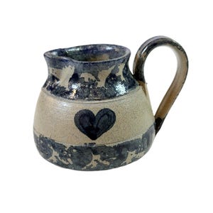 May include: A handmade ceramic pitcher with a heart design. The pitcher has a beige body with a dark blue rim and base. A black heart is painted on the front. The handle is a light brown color. This unique pitcher is perfect for serving beverages.