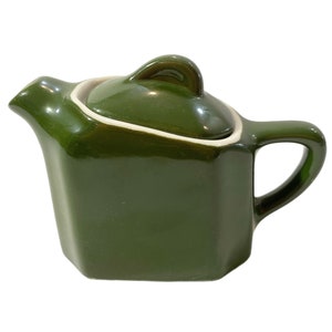 Puede incluir: Tetera pequeña de cerámica verde oscuro con tapa a juego. La tetera tiene un cuerpo cuadrado, un pico curvo y un asa. La tapa tiene un asa de bucle. Un borde blanco acentúa el borde de la tapa.