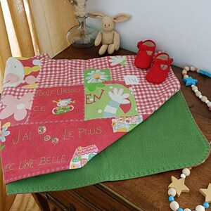 Peut inclure: Une couverture pour bébé rouge et verte avec un motif patchwork, des motifs floraux et vichy. La couverture porte du texte français et est associée à des chaussures de bébé rouges et une guirlande de perles en bois en forme d'étoile. Un lapin en peluche est à proximité.