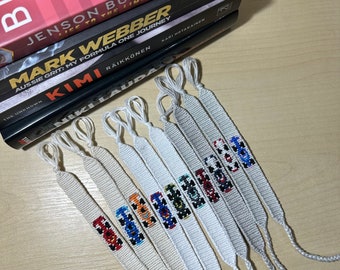 May include: A collection of white woven friendship bracelets, each featuring a different embroidered race car design. The bracelets are arranged near a stack of Formula 1 racing books, including "Jenson Button: Life to the Limit" and "Mark Webber: Aussie Grit."